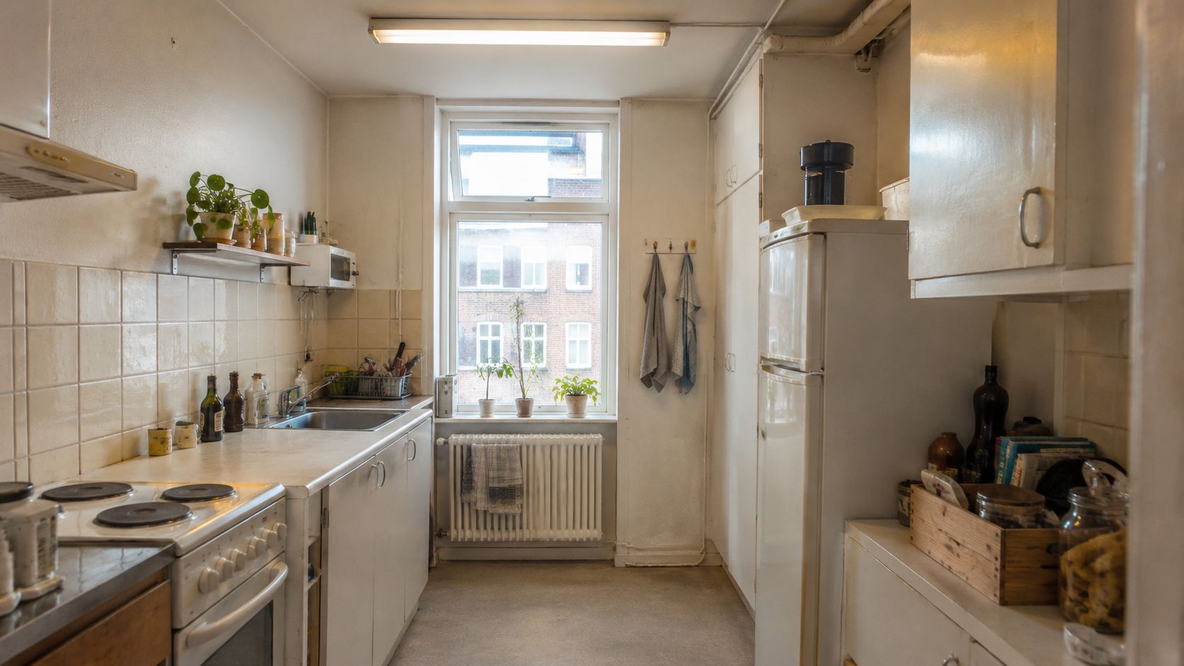 Et lyst og enkelt køkken i en lejlighed med hvide elementer, fliser og udsigt til en baggård.